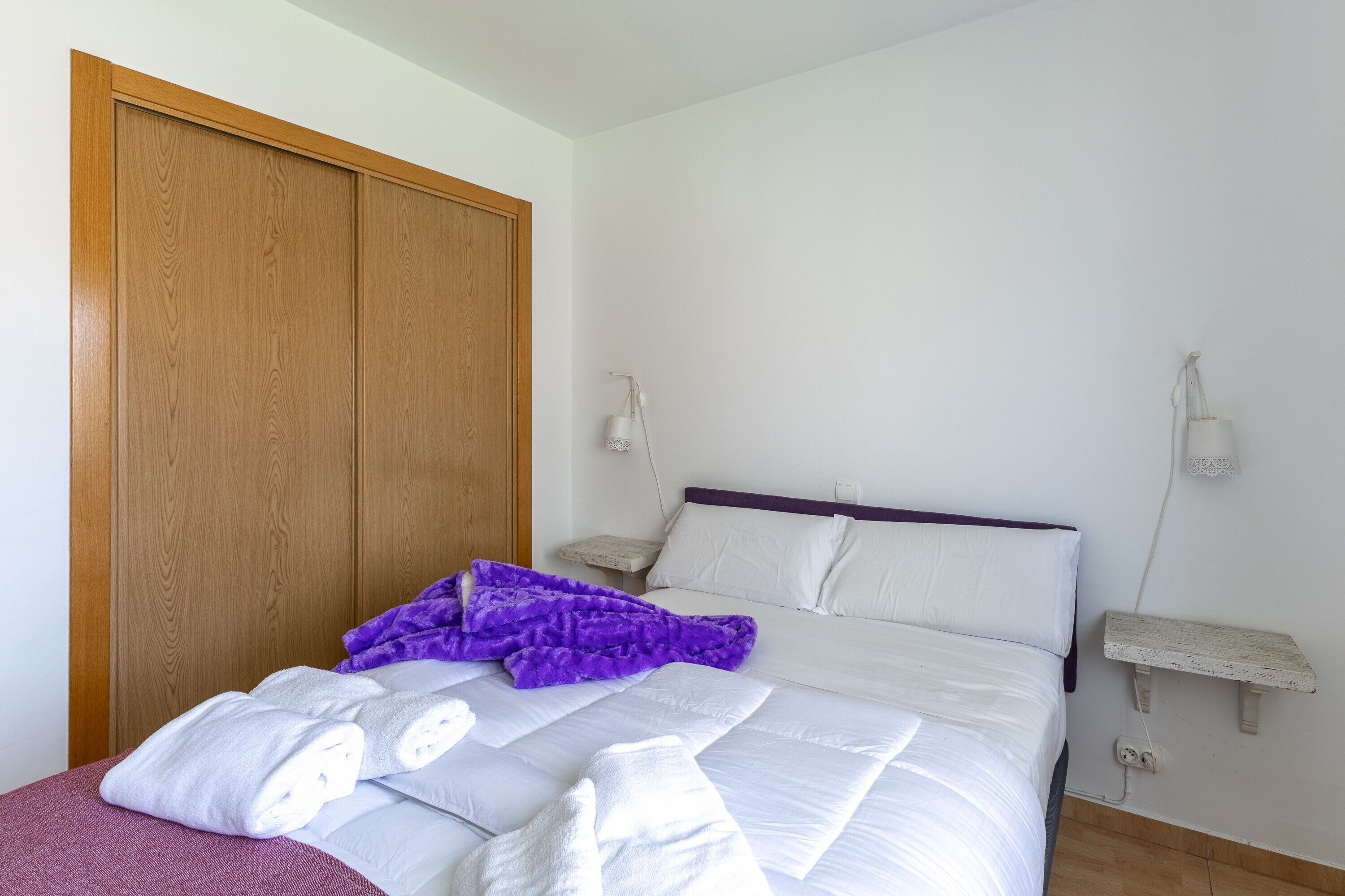Un dormitorio con un armario de madera, una cama con ropa de cama blanca, un edredón morado y dos mesitas de noche con lámparas.