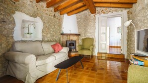 A cozy living room with a stone wall, a sofa, a wooden ceiling, a fireplace, a chair, and a TV. - Can Miquel: Charm and Pool in Pollença (Pollensa)