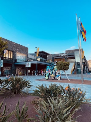 A modern building with a 'Hola!' sign, a 'lefties' store, and a large colorful sculpture in the plaza.
