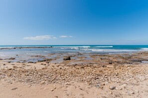 Una spiaggia con rocce e ciottoli sparsi, cielo azzurro intenso e onde oceaniche calme.