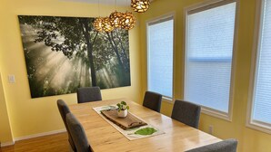 Un coin repas avec une table en bois, six chaises grises, un grand tableau représentant des arbres et une plante en pot posée sur la table.