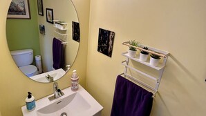 Une salle de bain avec un lavabo blanc, un miroir avec éclairage intégré, une serviette violette et une étagère avec des plantes.