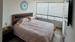 A bedroom with a bed, a bedside table, a wall-mounted TV, and a window with blinds.