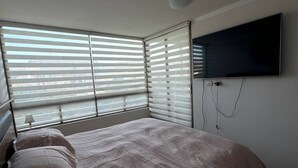 A bedroom with a bed, a TV mounted on the wall, and large windows with blinds.