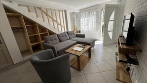 A modern living room with a grey sectional sofa, a wooden coffee table, and a TV on a wooden stand. There is a staircase with wooden railings leading to a loft area.