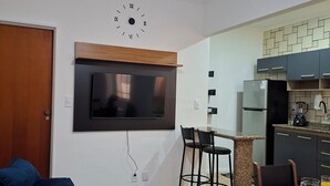 Een moderne woonkamer met een flatscreen televisie, een plafondventilator en een eethoek, met een keuken op de achtergrond.