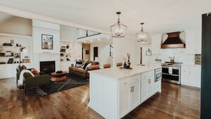 A spacious living area with a fireplace, a large island with a sink, and a kitchen with white cabinets and stainless steel appliances. - Heated Pool, Hot Tub, Game Room, EV charger, Sleeps 20 (Lawrenceburg)