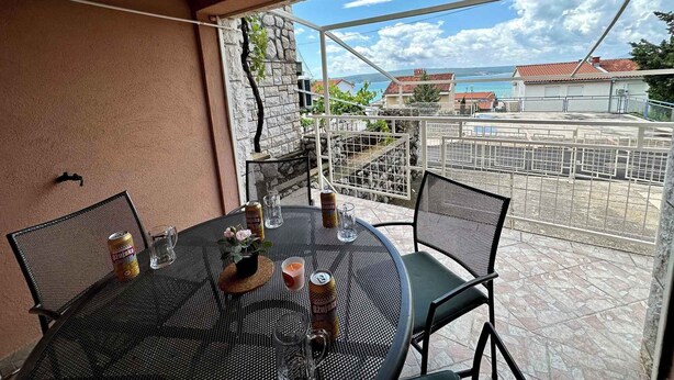 Una mesa redonda con sillas, un vaso de agua, una lata y una vela en un balcón con vistas a una zona costera con casas y un cielo despejado.