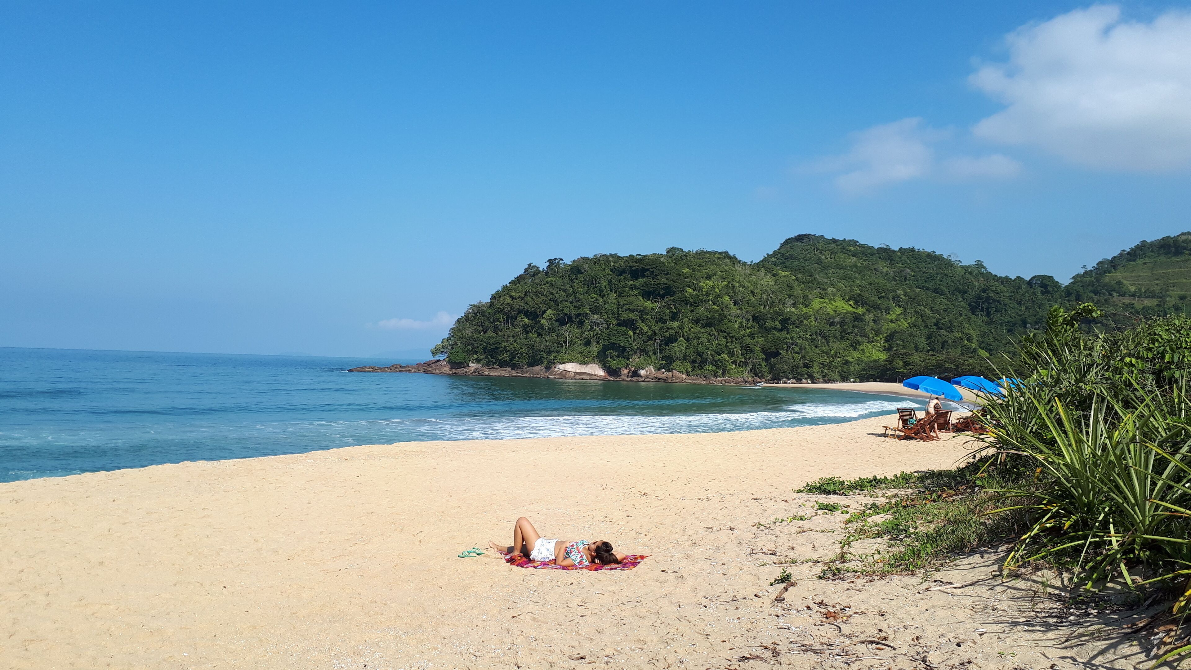 A beach with a person lying on a blanket, a blue umbrella, and a forested hill in the background.