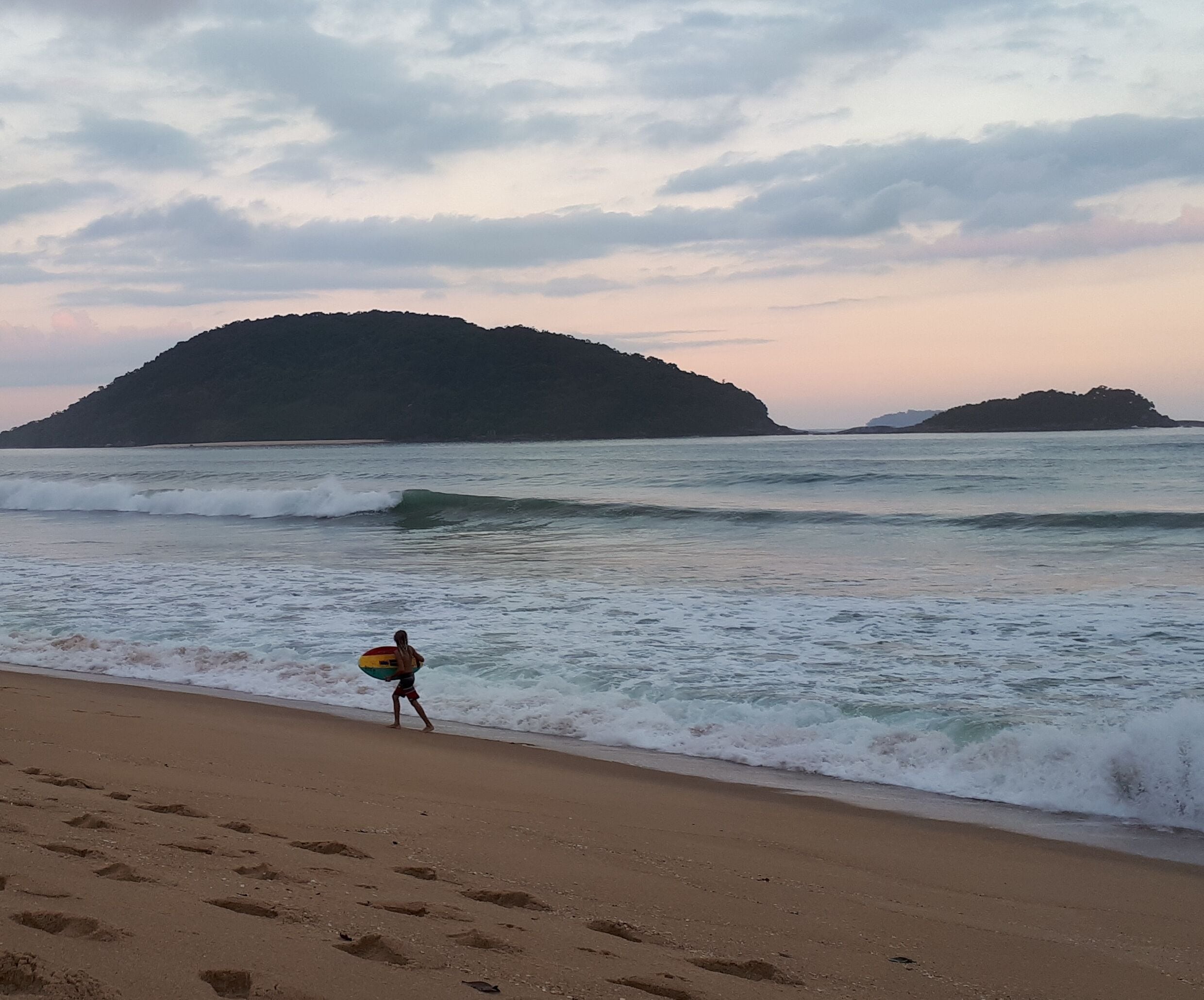 A beach with footprints leading to the water, a person carrying a surfboard, and distant islands under a cloudy sky.