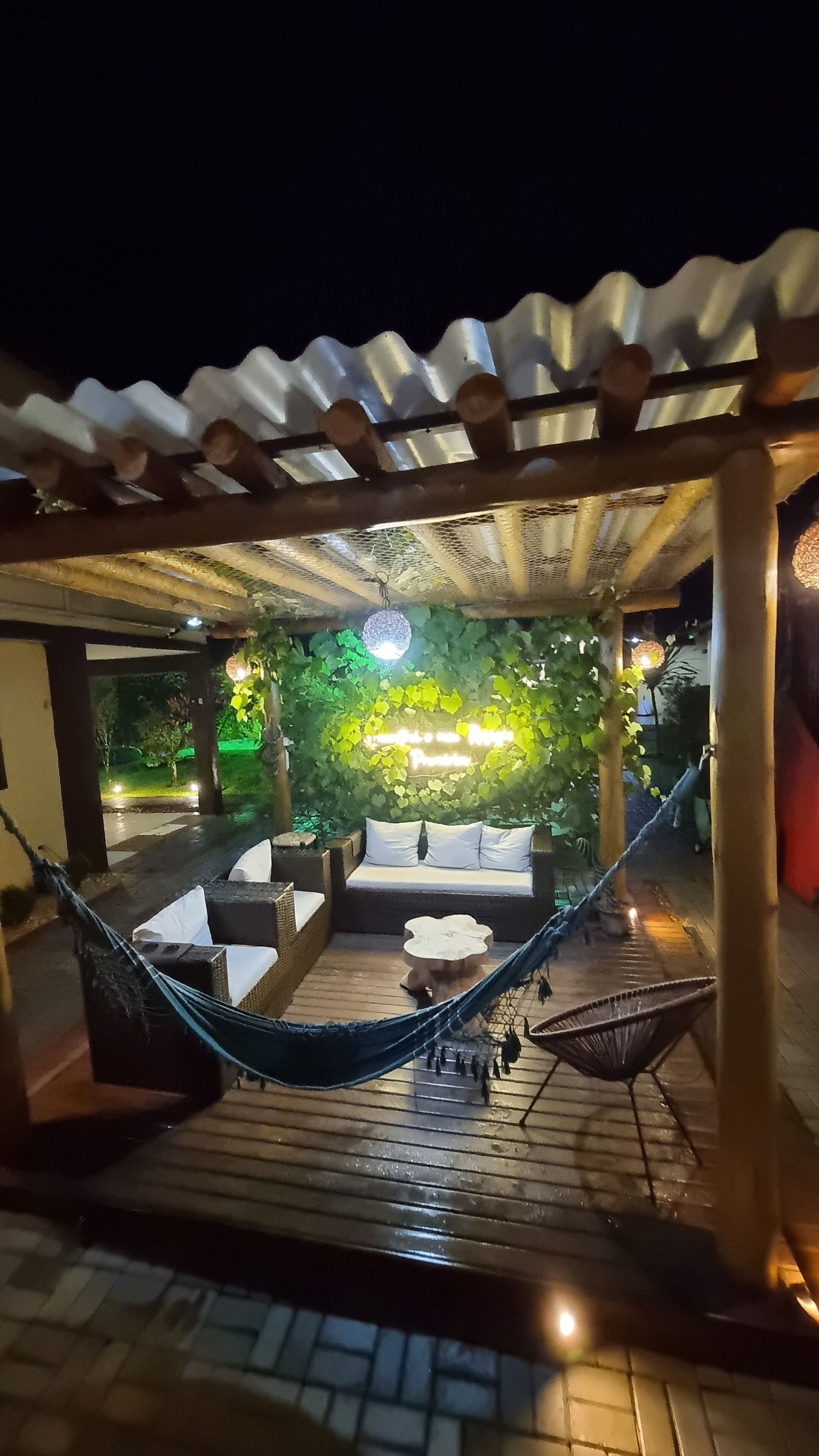 Uma área de estar ao ar livre com uma rede, móveis de madeira e um letreiro de neon ao fundo.
