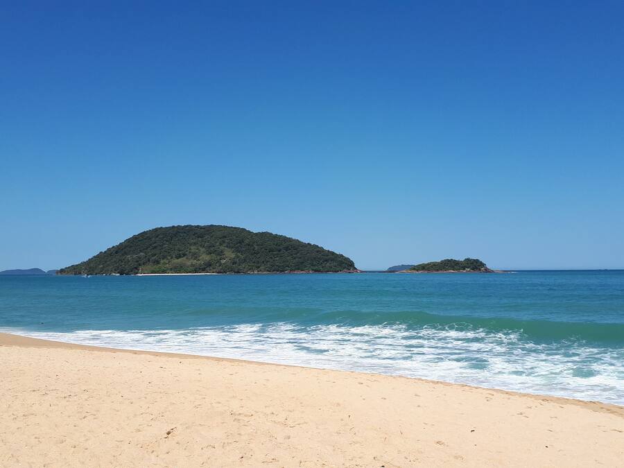 Una playa con aguas azules cristalinas, orilla arenosa y dos islas pequeñas a lo lejos.