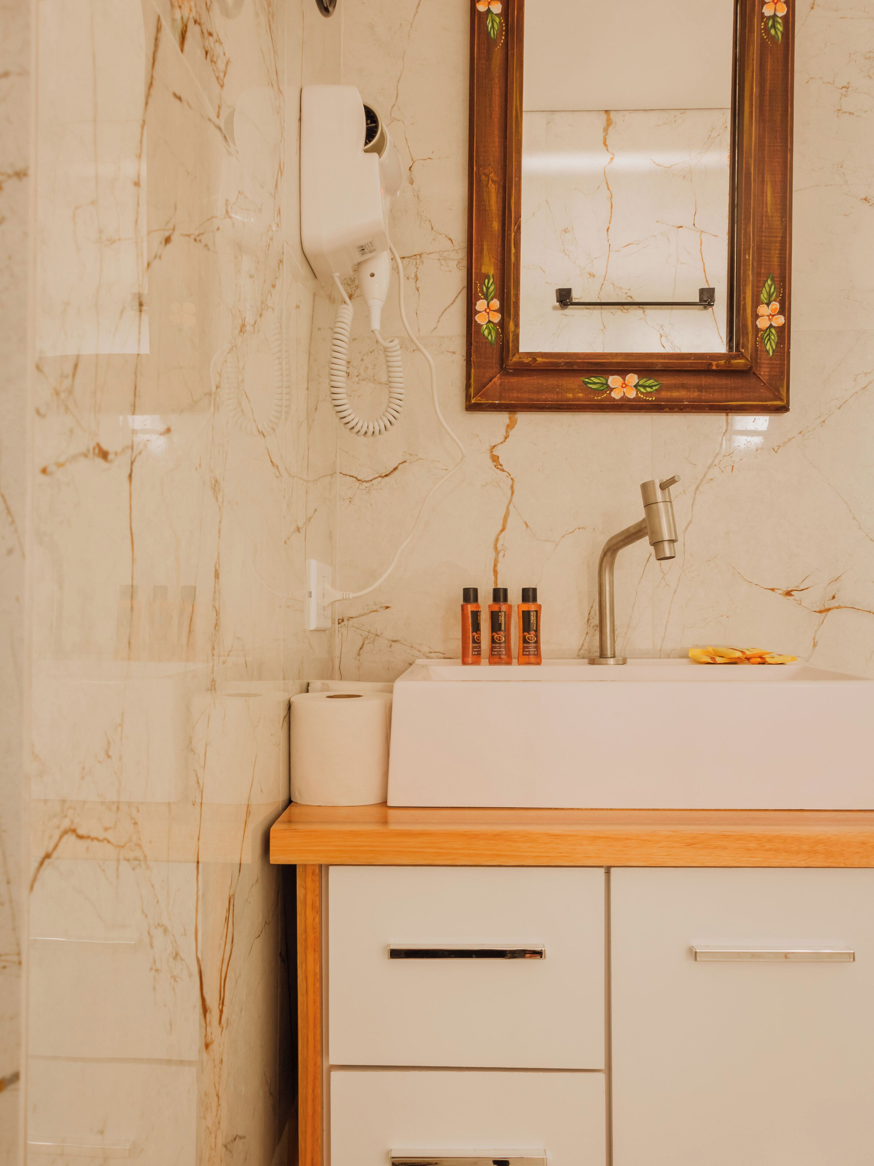 A bathroom with a marble wall, a wooden vanity, a sink, a mirror with floral decoration, and a hairdryer.