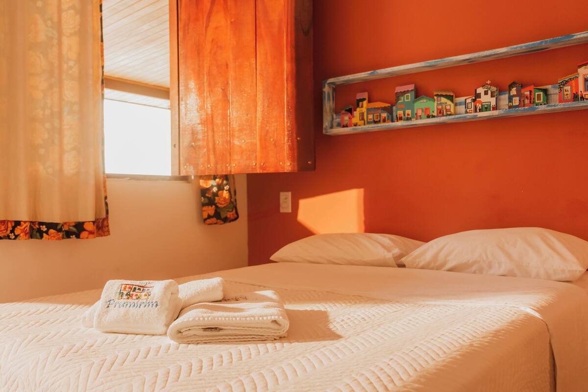 Un lit bien fait, avec du linge de lit blanc et une serviette pliée posée sur le dessus. Au-dessus du lit, on aperçoit une étagère avec des maisons miniatures colorées.