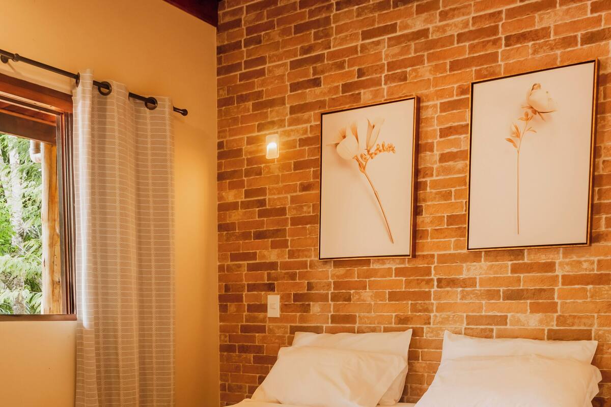 Un lit bien fait, avec du linge de lit blanc, un mur en briques et deux œuvres d’art florales encadrées au-dessus.