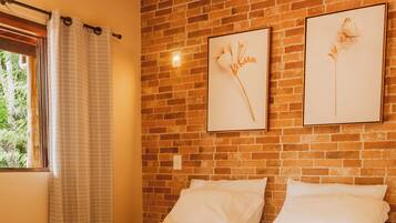 A neatly made bed with white linens, a brick wall, and two framed floral artworks above it.