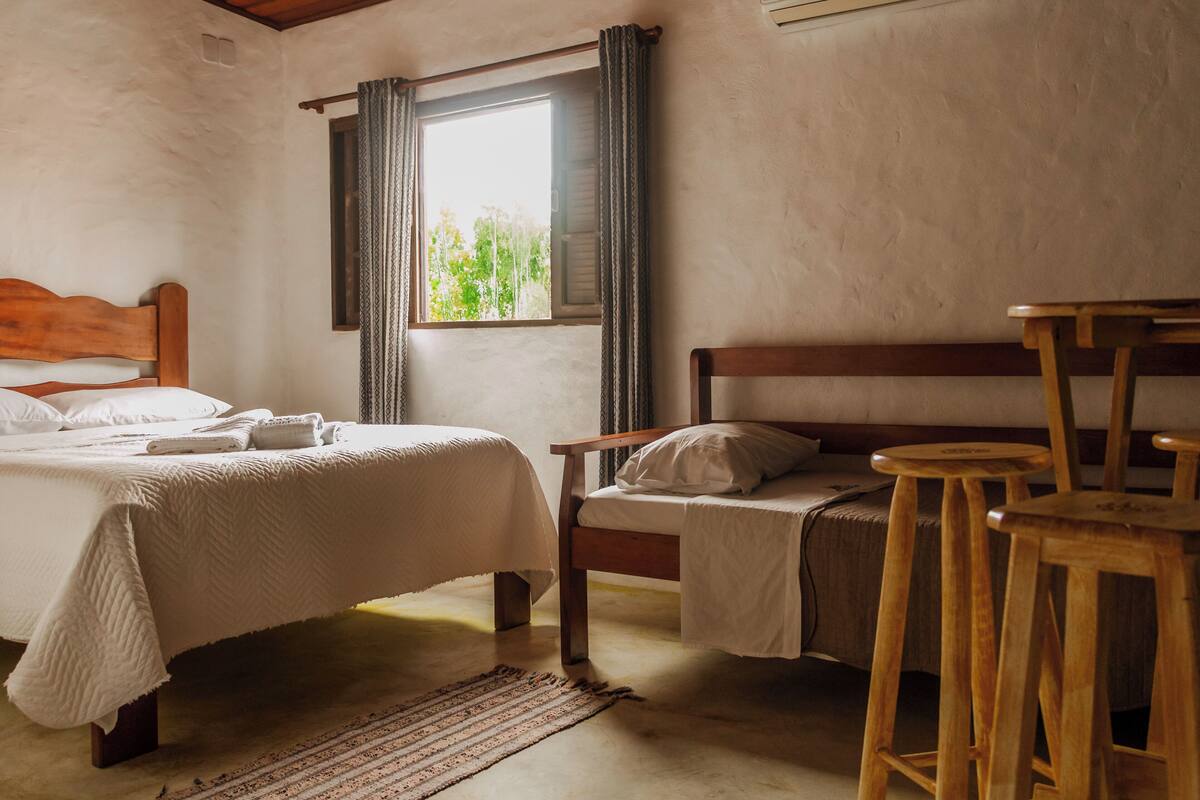 Une chambre avec un lit en bois, un plus petit lit, un banc en bois et une fenêtre avec des rideaux.