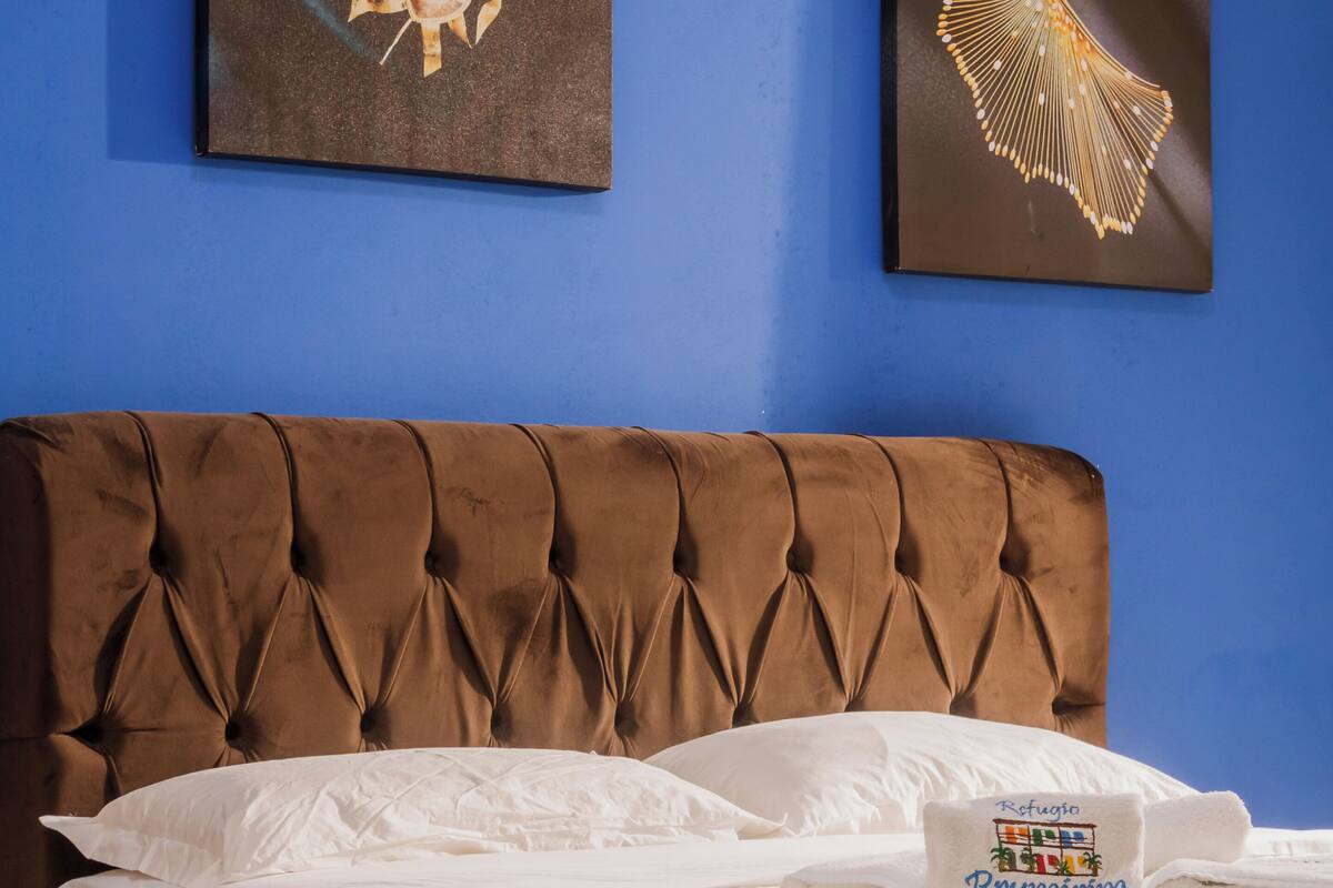 Un lit bien fait, avec des draps blancs et une tête de lit marron recouverte de tissu. Une serviette pliée, sur laquelle on peut lire le texte « Refugio Prumirim », est posée sur le lit.