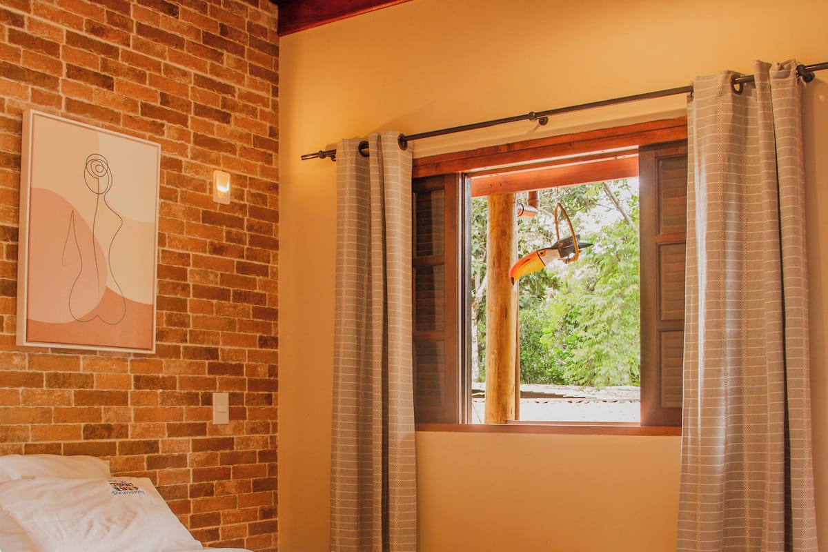 Une chambre avec un mur en briques, un lit avec une literie blanche, une fenêtre avec des rideaux et un luminaire suspendu.