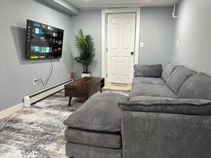A living room with a gray sectional sofa, a wooden coffee table, a potted plant, and a flat-screen TV mounted on the wall.