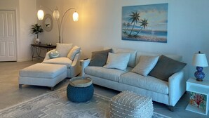 Een woonkamer met een bank, een poef en een schilderij van een strand.