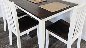 Une table à manger entourée de chaises blanches, une table en bois foncé et, au mur, un tableau encadré représentant une boutique de fleurs.