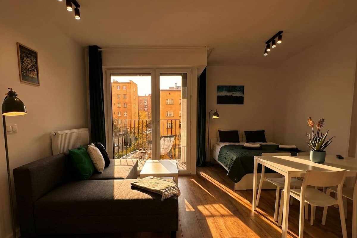 A cozy apartment with a sofa, a small dining table, a bed, and a balcony with a view of buildings.