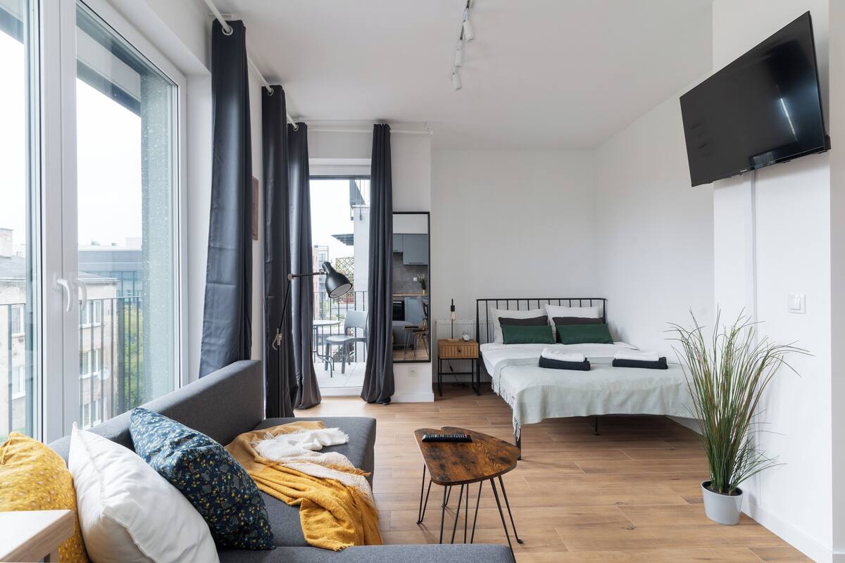A modern, well-lit room with a large window, a sofa, a bed, a TV, and a potted plant.