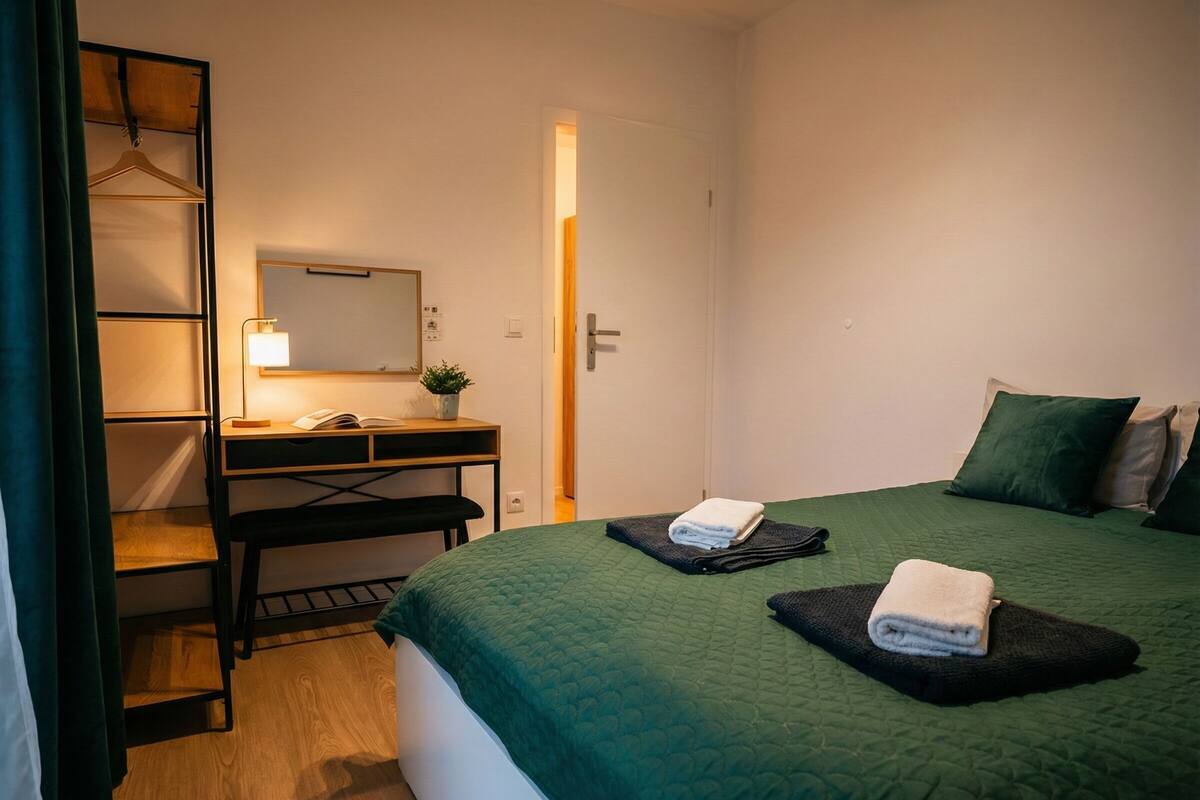 A hotel room with a bed, a desk, a chair, a hanging wardrobe, and a small potted plant.