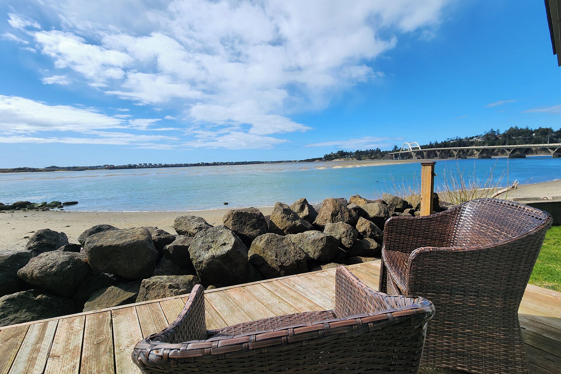 Une terrasse en bois, agrémentée de chaises en osier, offre une vue sur une plage parsemée de gros rochers et sur une étendue d’eau dégagée.