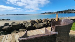 Een houten terras met rieten stoelen, met uitzicht op een strand met grote rotsen en helder uitzicht op het water.