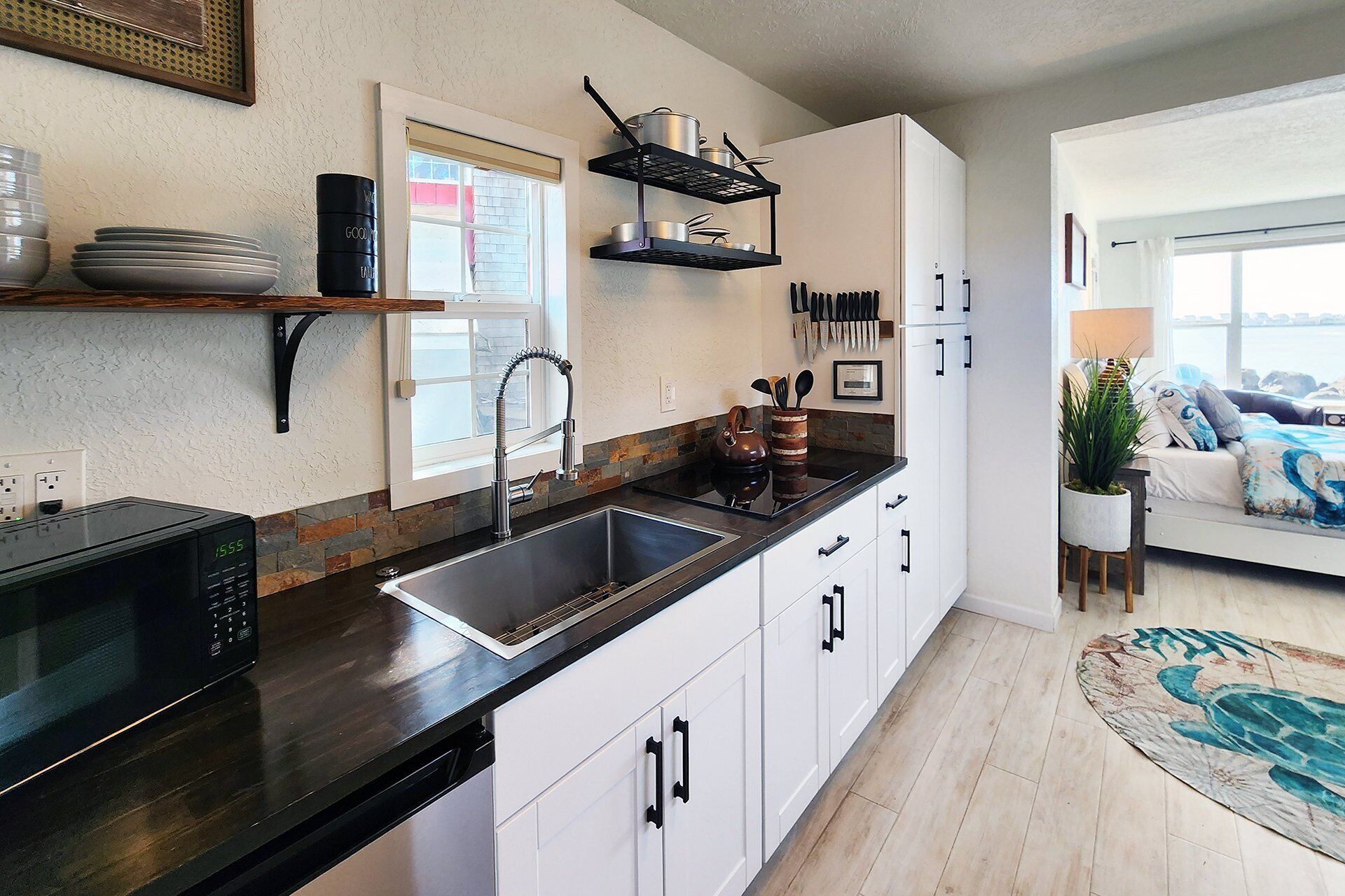 A modern kitchen with a black countertop, white cabinets, and a built-in sink. There is a window with blinds, a hanging pot rack, and a potted plant.