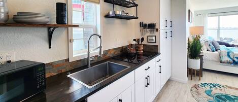 A modern kitchen with a black countertop, white cabinets, and a built-in sink. There is a window with blinds, a hanging pot rack, and a potted plant.