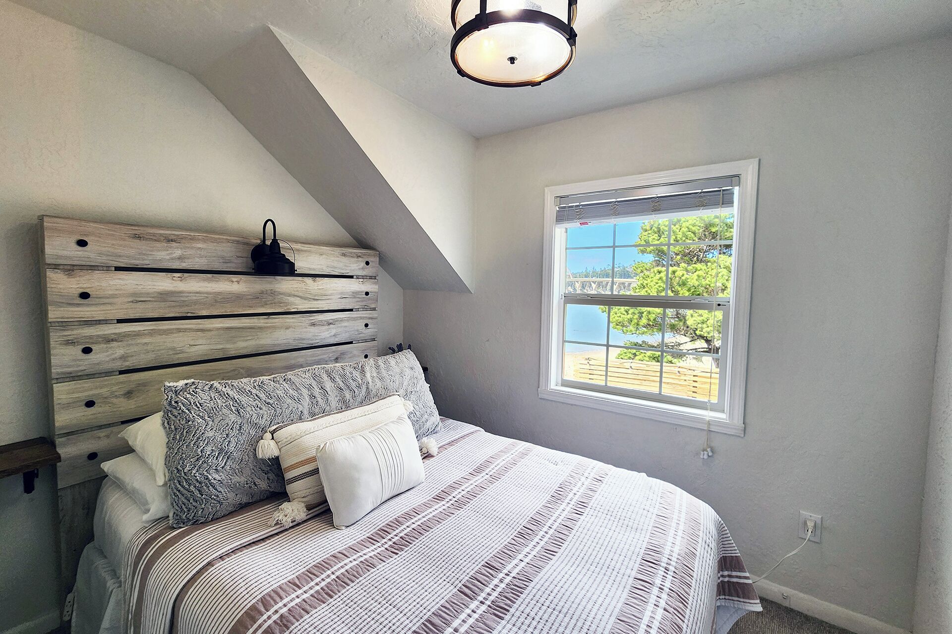 A bedroom with a sloped ceiling, a wooden headboard, a bed with striped bedding, and a window with a view of trees.
