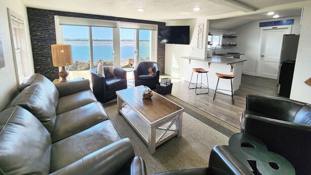 Een moderne woonkamer met een leren bank, een salontafel en een televisie. Er is een keukenhoek met een bar en een bord waarop 'BEACH' staat.