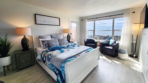 Een slaapkamer met een groot bed, twee bedlampen, uitzicht op zee, een stoel en een bijzettafeltje met een plant.