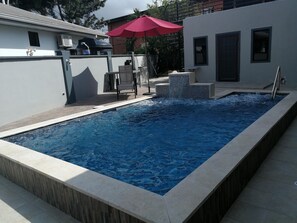 Une piscine rectangulaire bordée de carreaux, un parasol rouge et une chaise.