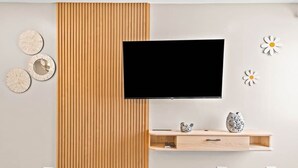 A modern living room with a flat-screen TV mounted on the wall, wooden paneling, a wooden shelf with decorative items, and wall art. - Peaceful Modern HNL Getaway (Honolulu)