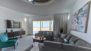 Ein modernes Wohnzimmer mit einem großen grauen Sofa, petrolfarbenen Sesseln und einem Couchtisch. Ein Flachbildfernseher ist an der Wand montiert, und ein großes abstraktes Gemälde ist sichtbar.