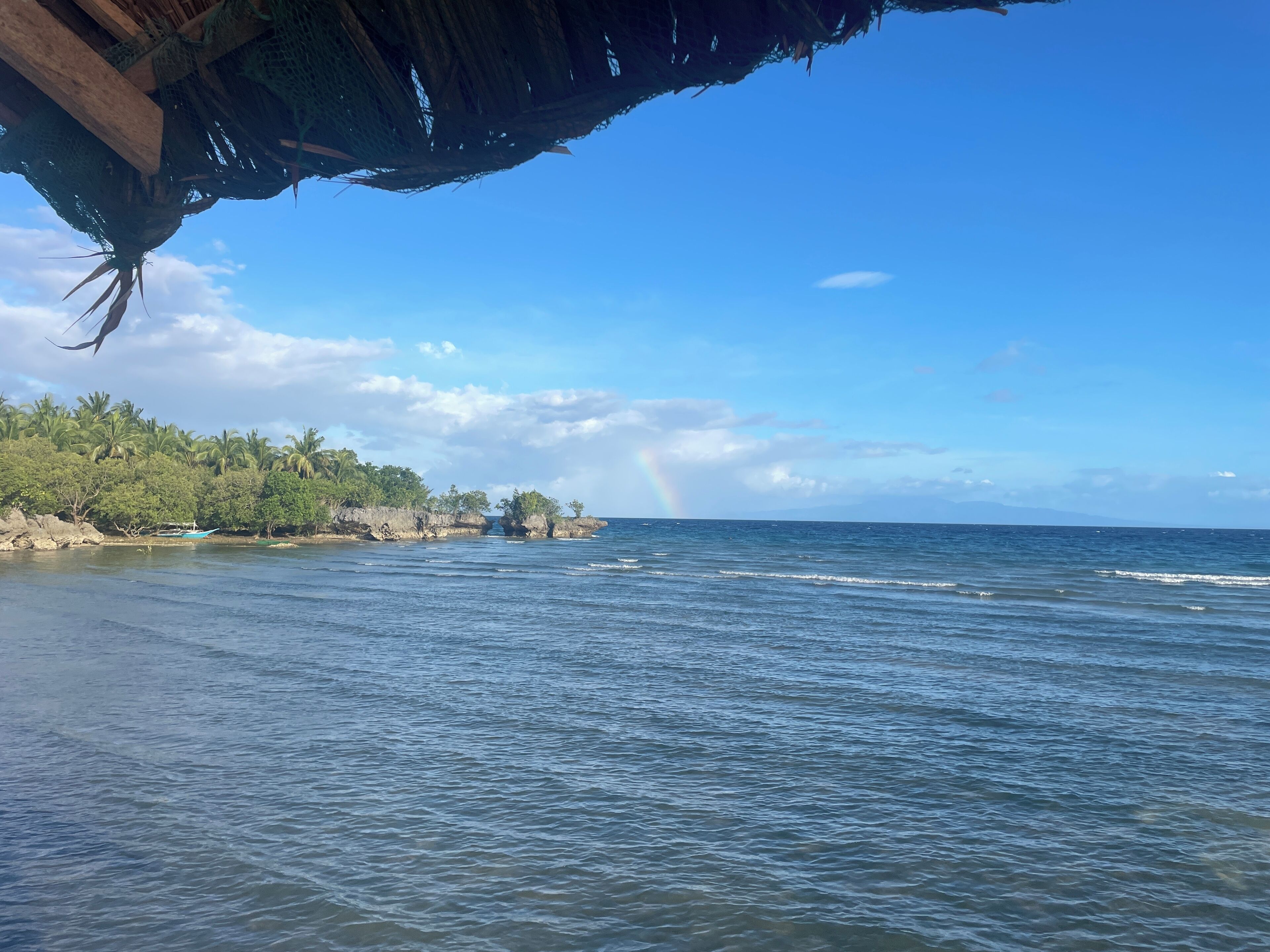 A beach with a thatched roof structure, calm sea, and a rainbow in the distance.