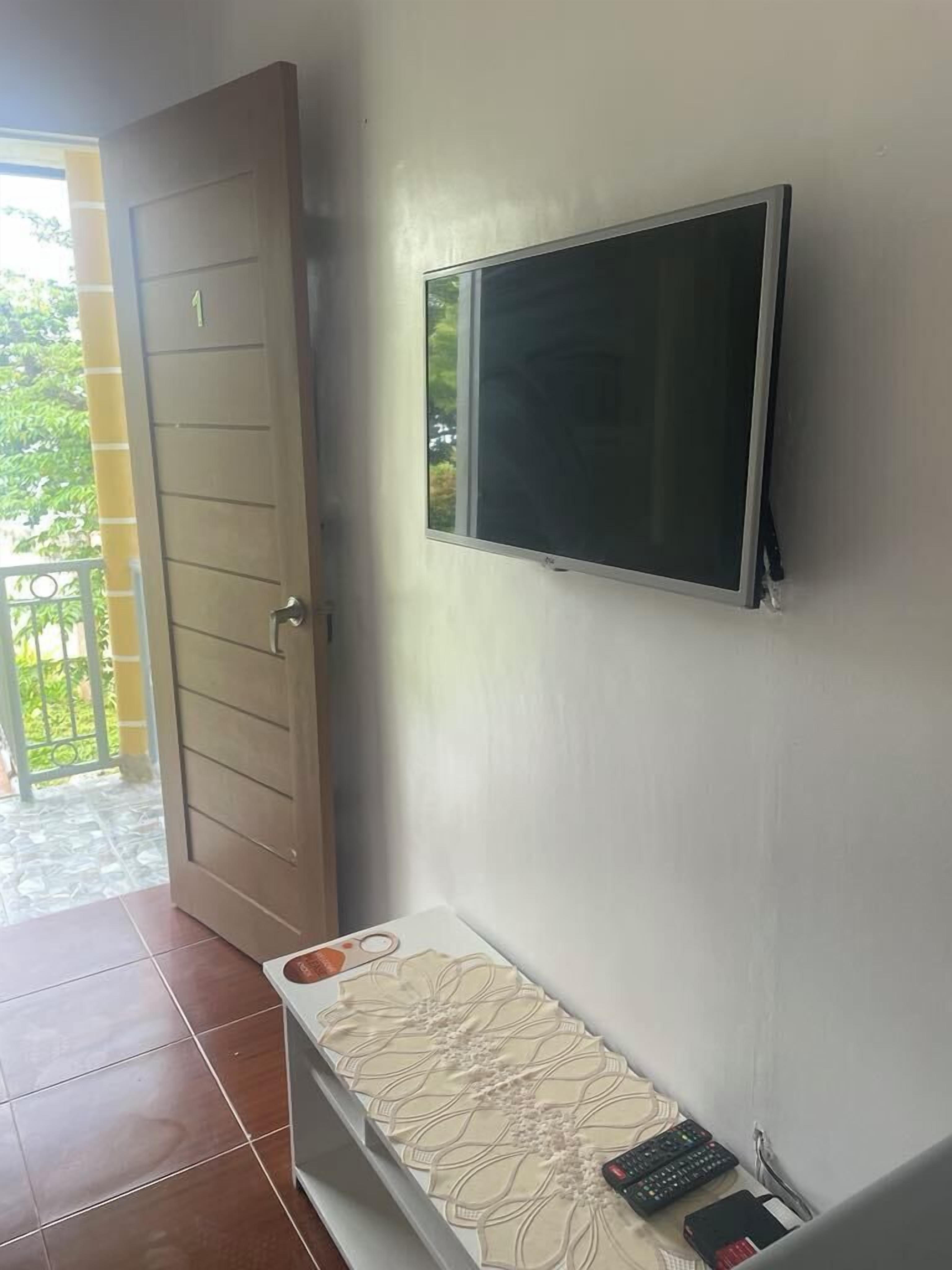 A room with a door, a wall-mounted TV, a small table with a decorative mat, and a remote control on the table.