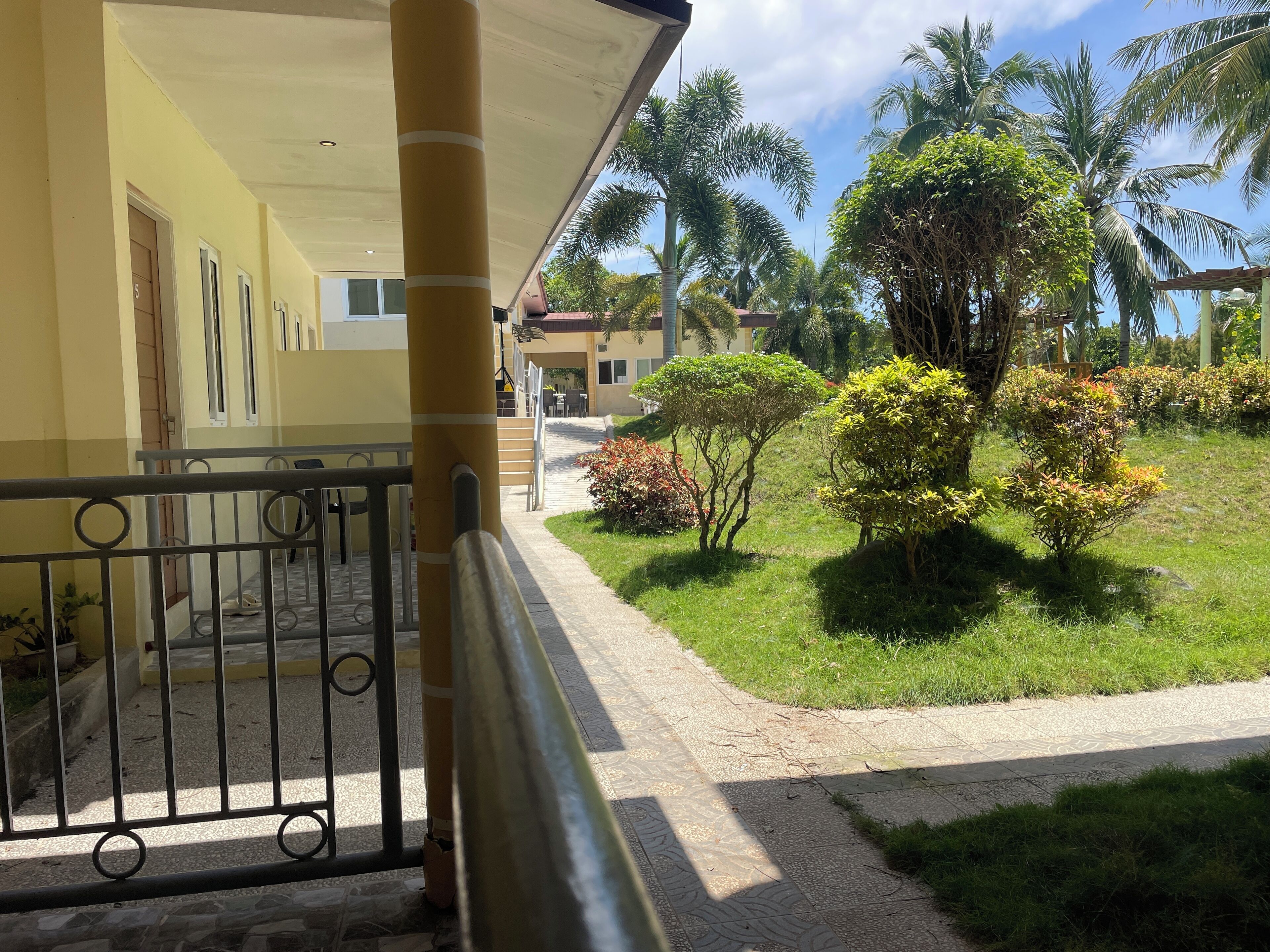 A yellow building with a covered balcony, a paved walkway, and palm trees in the background.