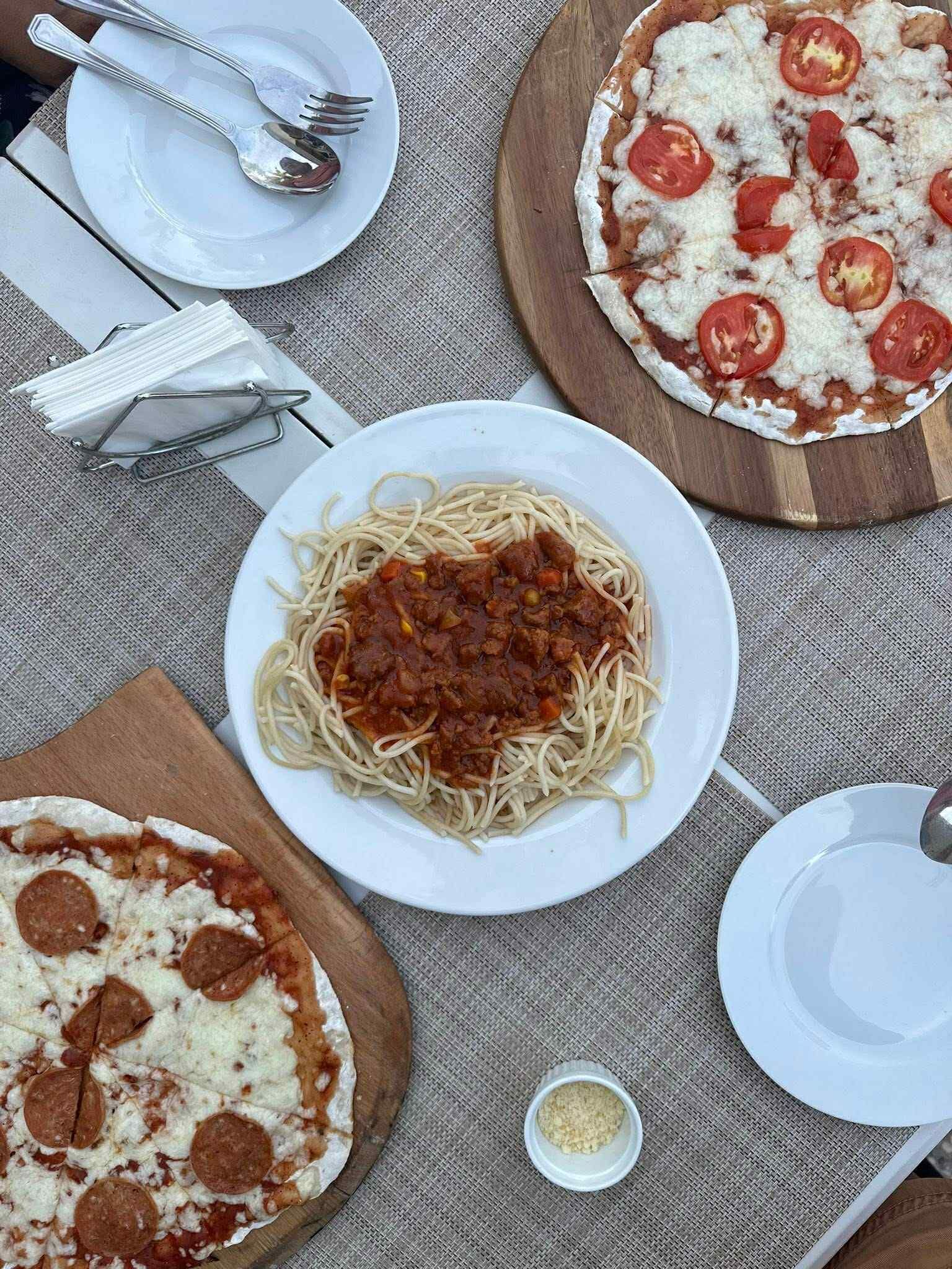 A table with three plates of food: spaghetti with tomato sauce, a pizza with tomatoes and cheese, and a pizza with pepperoni.