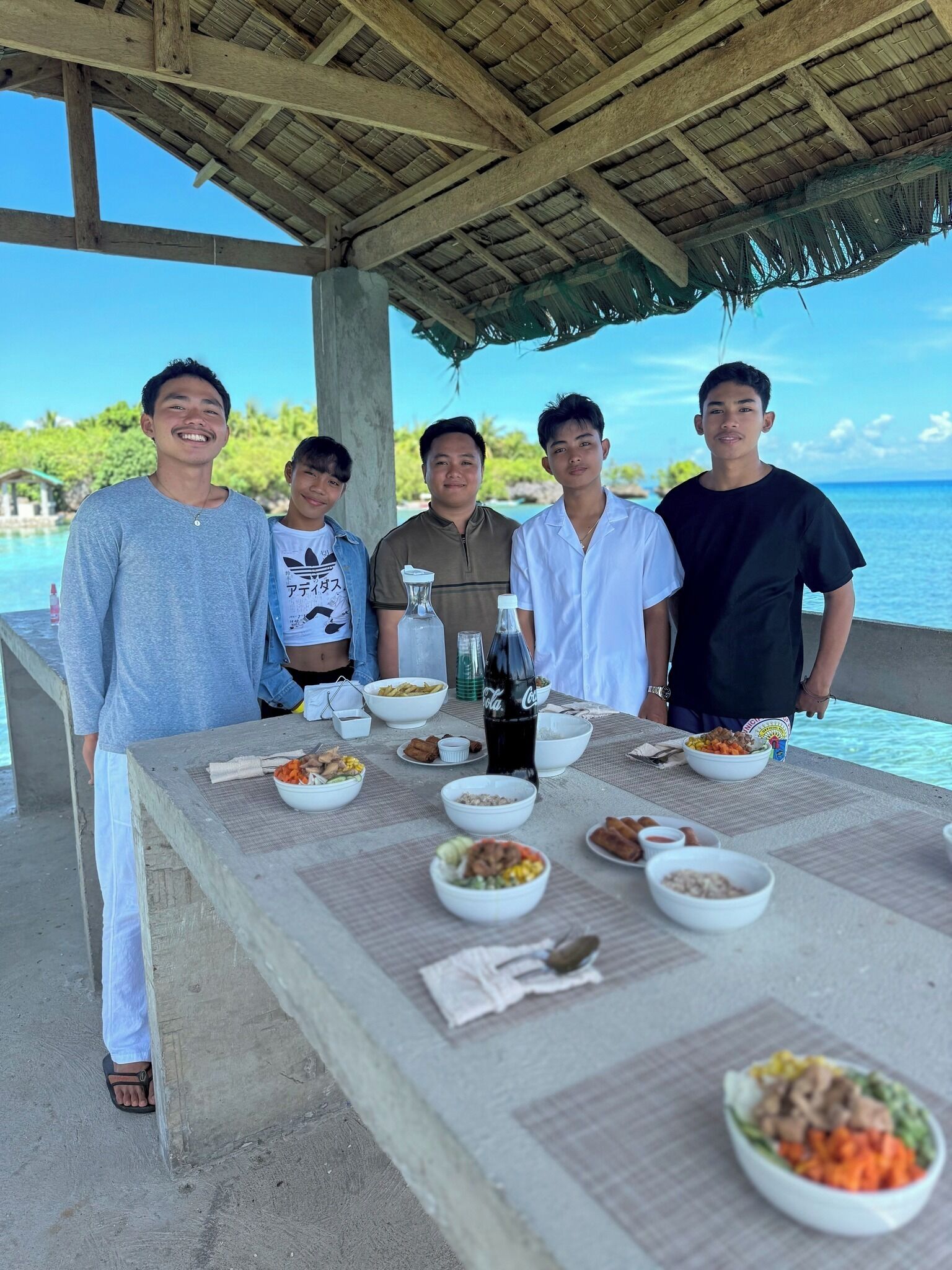 A group of people standing at a table with various dishes and a bottle of Coca-Cola, overlooking a clear blue sea.