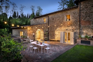 A stone building with a patio area featuring a table and chairs, surrounded by potted plants and a well-maintained lawn.