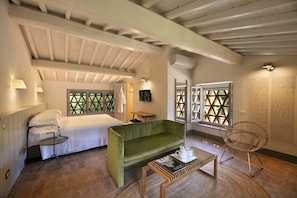 A cozy room with a bed, a green sofa, a small table, and a wicker chair. There are windows with a geometric pattern, a wall-mounted air conditioner, and a lamp on the wall.