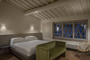 A bedroom with a bed, a green armchair, a wicker chair, a radiator, and a window with a lattice design.