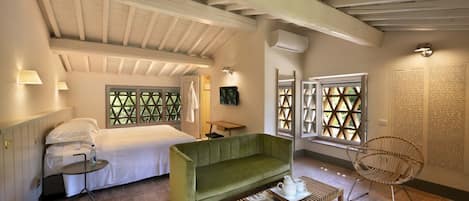 A cozy room with a bed, a green sofa, a small table, and a wicker chair. There are windows with a geometric pattern, a wall-mounted air conditioner, and a lamp on the wall.