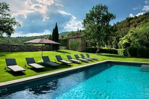 A swimming pool with lounge chairs and an umbrella, surrounded by a well-manicured lawn and trees.