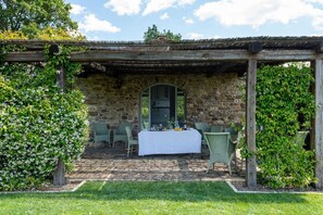 An outdoor dining area with a stone wall, wooden pergola, and green chairs.